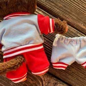 May include: A brown plush monkey doll wearing a light blue and red outfit. The jacket and shorts have white stripes. The monkey's tail is visible. The shorts are separate from the doll.