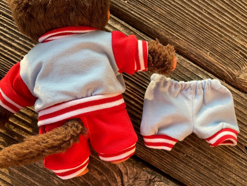 May include: A brown plush monkey doll wearing a light blue and red outfit. The jacket and shorts have white stripes. The monkey's tail is visible. The shorts are separate from the doll.
