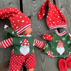 May include: A red and white gnome-themed outfit for a doll. The outfit includes a red hat with white polka dots, a green and white striped shirt with a gnome design, red trousers with white polka dots, and a pair of red slippers.