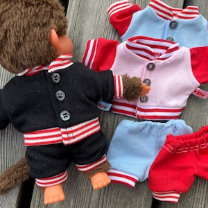 May include: A monkey doll wearing a black jacket and shorts with red and white striped trim. Additional jackets in pink and light blue, and blue and red shorts are also visible. The doll has brown fur and orange hands and feet.