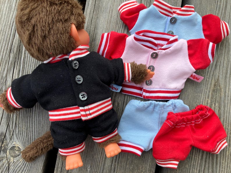 May include: A monkey doll wearing a black jacket and shorts with red and white striped trim. Additional jackets in pink and light blue, and blue and red shorts are also visible. The doll has brown fur and orange hands and feet.