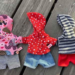 May include: Three handmade doll outfits with hoodies. The first outfit is pink with a black and white elephant print, the second is red with white polka dots, and the third is blue and gray striped. Each outfit has a small label that says "Kipu".