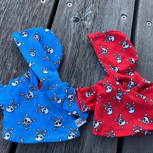 May include: Two hooded sweatshirts with skull and crossbones patterns. One is blue with white skulls and the other is red with white skulls. Both have a tag that says "kipu".
