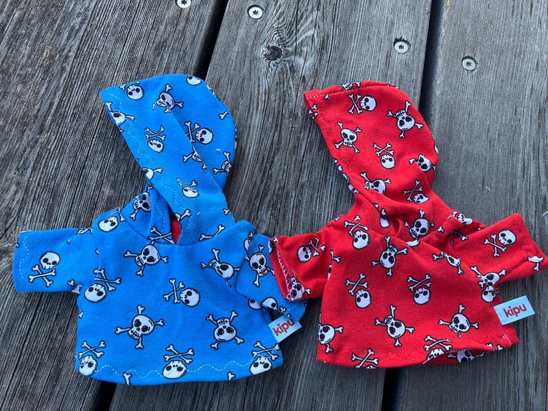 May include: Two hooded sweatshirts with skull and crossbones patterns. One is blue with white skulls and the other is red with white skulls. Both have a tag that says "kipu".