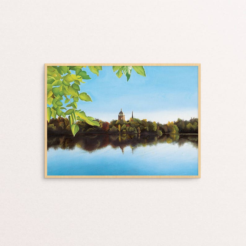 May include: A painting of a lake with a small island in the distance. The island has a building with a spire on it. The lake is surrounded by trees and the sky is blue. The painting is framed in wood.