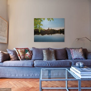 May include: A gray couch with pillows in a living room setting. The couch is in front of a wall with a framed print of a lake scene with trees and a building in the distance. The print is above a glass coffee table with books stacked on top.