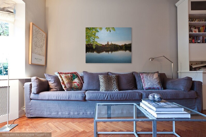 May include: A gray couch with pillows in a living room setting. The couch is in front of a wall with a framed print of a lake scene with trees and a building in the distance. The print is above a glass coffee table with books stacked on top.