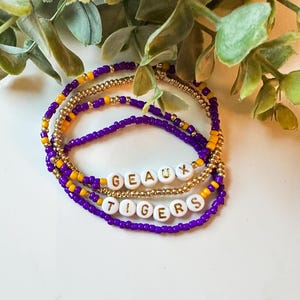 May include: A set of four beaded bracelets with the words "Geaux Tigers" spelled out in gold letters on white beads. The bracelets are made with purple and gold beads.