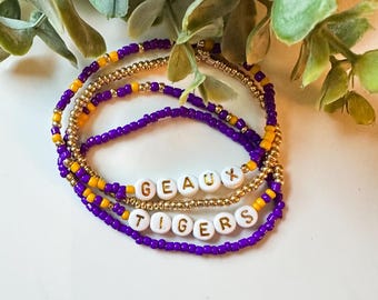 May include: A set of four beaded bracelets with the words "Geaux Tigers" spelled out in gold letters on white beads. The bracelets are made with purple and gold beads.