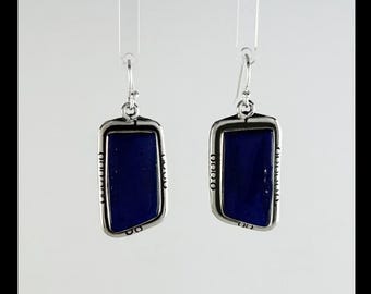 May include: A pair of silver-toned rectangular earrings with a deep blue, possibly lapis lazuli, center. The earrings have a decorative border and a hook closure. The background is a plain white.