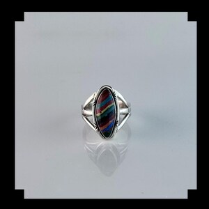 May include: A silver ring with an oval-shaped, multicolored stone inlay. The stone features a striped pattern in shades of red, blue, green, and yellow. The ring has a split shank design, adding visual interest to the jewelry.
