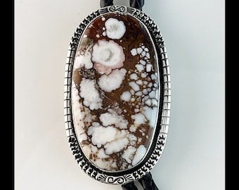 May include: A bolo tie with a silver oval setting featuring a white, brown, and pink agate stone. The bolo tie has a black braided cord.