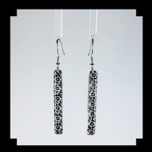 Op de afbeelding: Een paar zilveren oorhangers met een bloemenmotief. De oorhangers zijn lang en slank, met een delicate ketting en een klein bloemenbedeltje aan de onderkant.