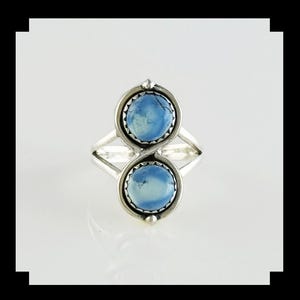 May include: A silver ring featuring two round, light blue stones with dark veining. Each stone is set in a serrated bezel mount. The band is split into two thin, polished silver wires that converge at the sides to support the vertical stone setting.