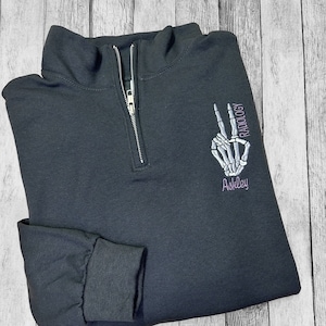 May include: Black quarter-zip sweatshirt with a skeleton hand making a peace sign and the text "RADIOLOGY Ashley" embroidered on the right side.