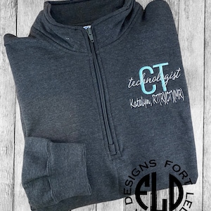 May include: A dark gray fleece pullover with a zipper. The pullover has a monogrammed "CT" on the right side with the text "technologist" and "Katelyn, RT(R)Y(CT)(MR)" below. The left sleeve has a black circle with the letters "FLD" inside and the text "BETTER" and "DESIGNS FORT" around the circle.