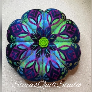 May include: A round, fabric pincushion featuring a vibrant mandala-inspired pattern in shades of purple, blue, and lime green. The center is accented with a small, lime green rose-shaped button. The text StaciesQuiltStudio is visible at the bottom.