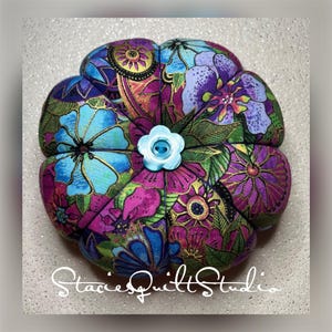 May include: A colorful floral pincushion with a blue button in the center. The fabric is a mix of pink, purple, blue, green, and gold.