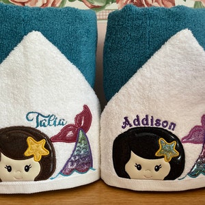 May include: Two white hooded towels with embroidered mermaid designs. The towels have teal blue trim and feature the names "Talia" and "Addison" in purple and pink.