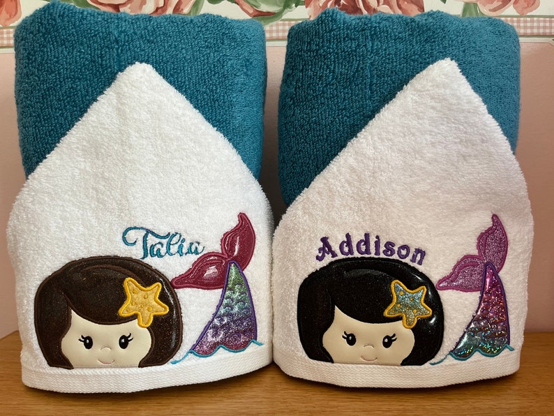 May include: Two white hooded towels with embroidered mermaid designs. The towels have teal blue trim and feature the names "Talia" and "Addison" in purple and pink.