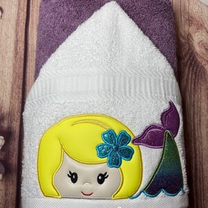 May include: A white hooded towel with an embroidered mermaid design. The mermaid has blonde hair, a blue flower, and a purple and green tail. The towel has the text "Stacie's Quilt Studio" at the bottom.