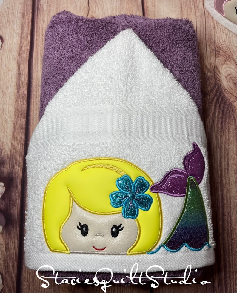 May include: A white hooded towel with an embroidered mermaid design. The mermaid has blonde hair, a blue flower, and a purple and green tail. The towel has the text "Stacie's Quilt Studio" at the bottom.