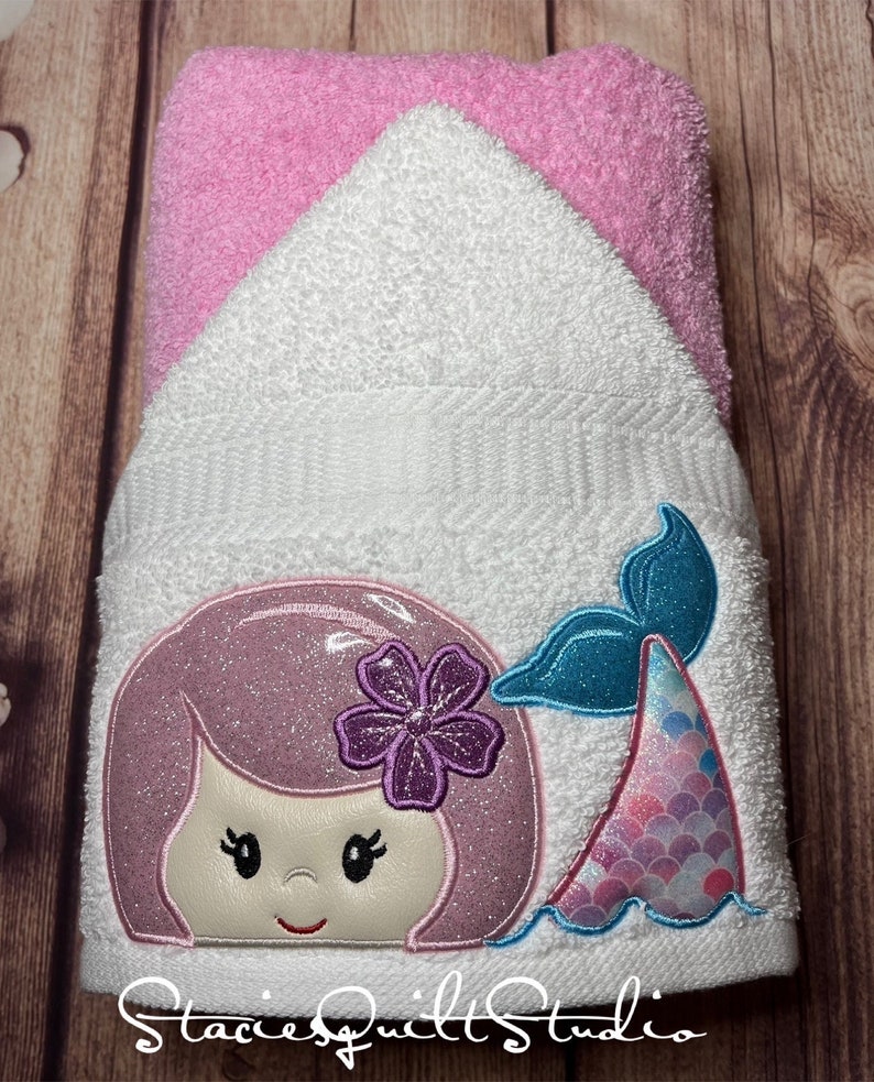 May include: A white hooded towel with a pink trim. The towel has an embroidered mermaid design with a purple flower in her hair and a blue tail with pink and blue scales.
