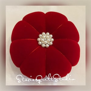 May include: A red velvet ring box with a flower-shaped design and a pearl embellishment in the center.