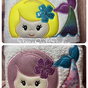 May include: Two embroidered towels with a mermaid design. The top towel features a yellow-haired mermaid with a blue flower and a purple tail. The bottom towel features a pink-haired mermaid with a purple flower and a blue tail with pink scales.
