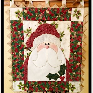 May include: A quilted wall hanging featuring a festive Santa Claus with a red hat, white beard, and a holly sprig. The background is a patchwork of green, red, and gold fabric with a Christmas theme. The bottom of the hanging has the text "Stacies Quilt Studio".