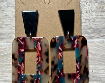 May include: A pair of dangle earrings with a black geometric top and a rectangular bottom. The bottom is made of a translucent material with a colorful swirl pattern.