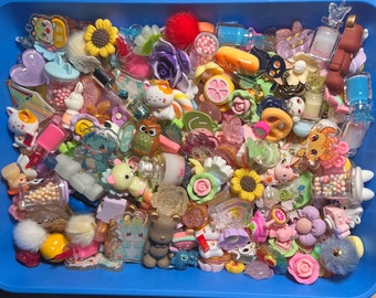 Puede incluir: Una colección de abalorios y baratijas coloridos en un recipiente de plástico. Los abalorios están hechos de varios materiales, incluyendo metal, plástico y vidrio. Algunos de los abalorios tienen forma de animales, flores y corazones. Otros tienen un diseño más abstracto. Los abalorios están dispuestos en un orden aleatorio, creando una exhibición visualmente atractiva y ecléctica.