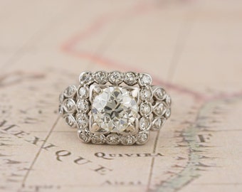 Puede incluir: Un anillo de compromiso de estilo vintage con un gran diamante redondo y claro en el centro. El diamante está rodeado por un halo cuadrado de diamantes más pequeños, con detalles adicionales de diamantes en la banda. El anillo está sobre un fondo claro.
