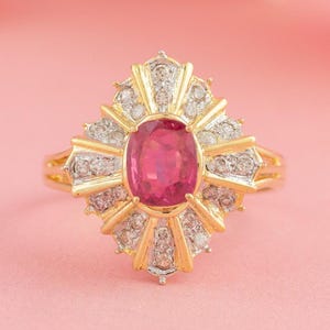 Puede incluir: Un anillo de oro con una gran piedra central ovalada de rubí, rodeada de diamantes más pequeños. El anillo tiene un diseño de rayos de sol y está sobre un fondo rosa con flores rosas borrosas.