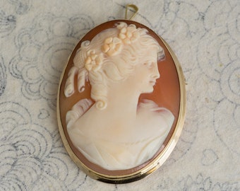 May include: A cameo brooch featuring a carved portrait of a woman with flowers in her hair, set in a gold tone oval frame.
