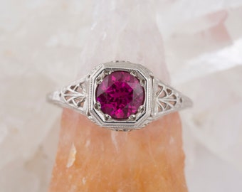 Puede incluir: Un anillo de compromiso vintage con una piedra preciosa rosa oscuro engastada en una banda de filigrana.