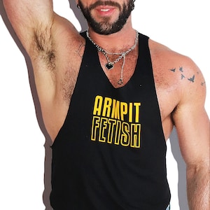 May include: A black sleeveless tank top with a yellow "ARMPIT FETISH" graphic print. The model is wearing the tank top with blue jeans.