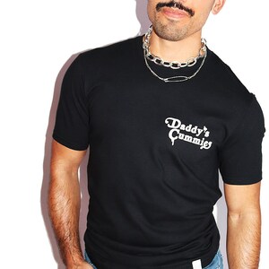 K&ouml;nnte beinhalten: Ein schwarzes T-Shirt mit dem Aufdruck "Daddy's Cummies" in wei&szlig; auf der Vorderseite. Die Person, die das Shirt tr&auml;gt, tr&auml;gt auch eine silberne Kettenkette und eine Sonnenbrille.