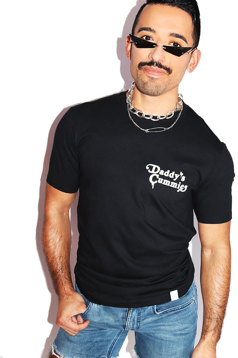 K&ouml;nnte beinhalten: Ein schwarzes T-Shirt mit dem Aufdruck "Daddy's Cummies" in wei&szlig; auf der Vorderseite. Die Person, die das Shirt tr&auml;gt, tr&auml;gt auch eine silberne Kettenkette und eine Sonnenbrille.