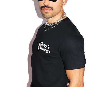 K&ouml;nnte beinhalten: Ein schwarzes T-Shirt mit einem wei&szlig;en Aufdruck, der "Daddy's Gummies" lautet. Die Person, die das Shirt tr&auml;gt, hat einen Schnurrbart und tr&auml;gt eine schwarze Sonnenbrille und eine silberne Kettenkette.