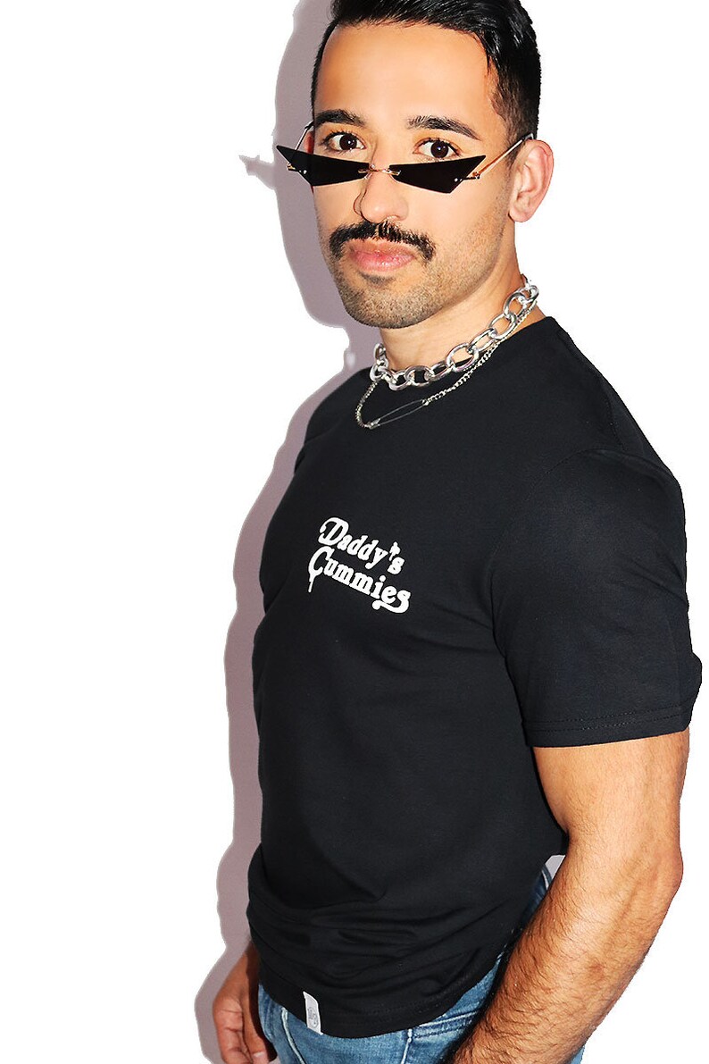 K&ouml;nnte beinhalten: Ein schwarzes T-Shirt mit einem wei&szlig;en Aufdruck, der "Daddy's Gummies" lautet. Die Person, die das Shirt tr&auml;gt, hat einen Schnurrbart und tr&auml;gt eine schwarze Sonnenbrille und eine silberne Kettenkette.