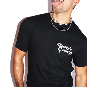 K&ouml;nnte beinhalten: Ein schwarzes T-Shirt mit einem wei&szlig;en Aufdruck, der "Daddy's Cummie's" lautet. Die Person, die das Shirt tr&auml;gt, tr&auml;gt eine silberne Kettenkette und eine Sonnenbrille.