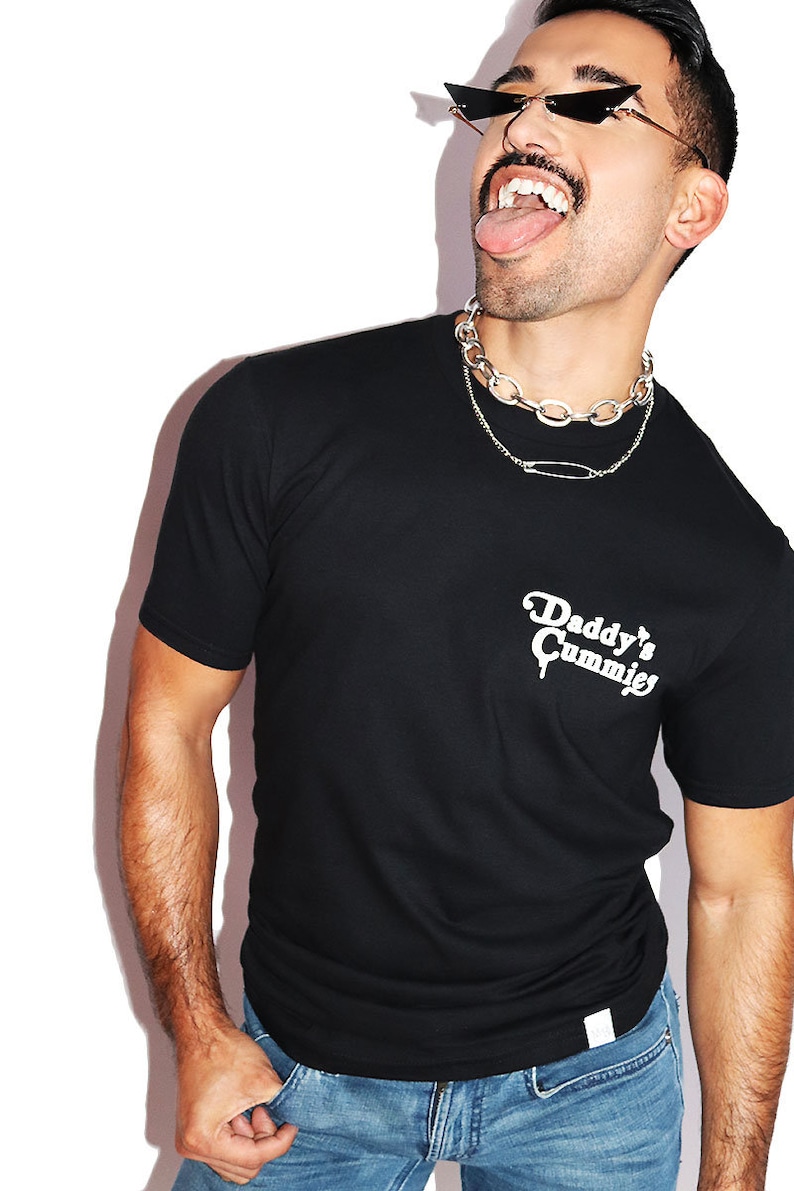 K&ouml;nnte beinhalten: Ein schwarzes T-Shirt mit einem wei&szlig;en Aufdruck, der "Daddy's Cummie's" lautet. Die Person, die das Shirt tr&auml;gt, tr&auml;gt eine silberne Kettenkette und eine Sonnenbrille.