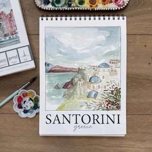 May include: A watercolor sketch of Santorini, Greece, on a white spiral-bound sketchbook. The artwork depicts the iconic blue-domed churches and coastal scenery. The word "SANTORINI" is printed in bold black letters, with "greece" below in cursive.