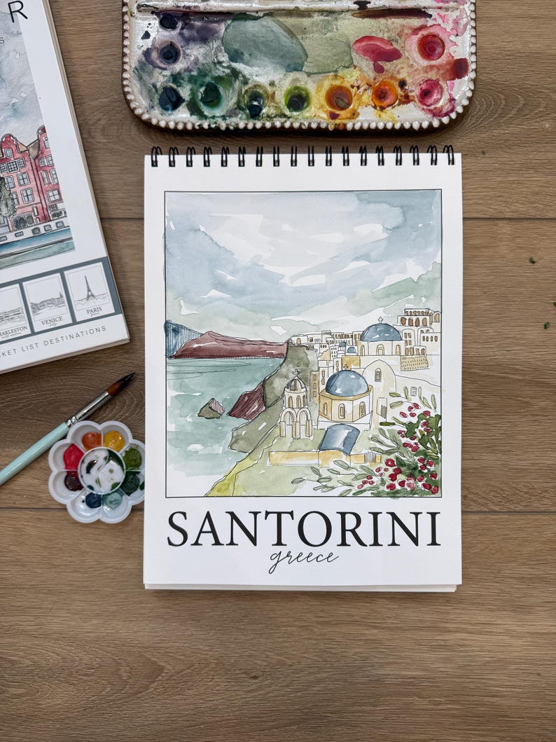 May include: A watercolor sketch of Santorini, Greece, on a white spiral-bound sketchbook. The artwork depicts the iconic blue-domed churches and coastal scenery. The word "SANTORINI" is printed in bold black letters, with "greece" below in cursive.