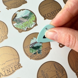 May include: A collection of brown paper circle stickers with illustrations of various national parks. The stickers feature text such as "Hawaii Volcanoes", "Kings Canyon", "New River Gorge", "Mount Rainier", "KOBUK VALLEY", and "SAGUARO".