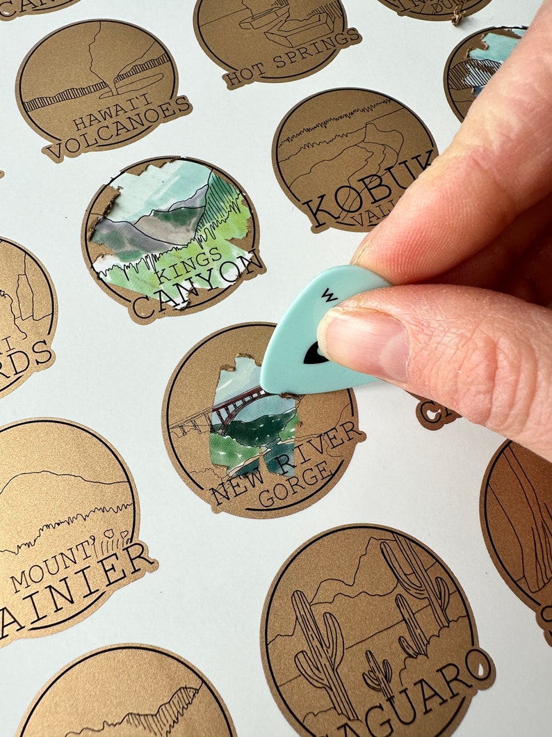 May include: A collection of brown paper circle stickers with illustrations of various national parks. The stickers feature text such as "Hawaii Volcanoes", "Kings Canyon", "New River Gorge", "Mount Rainier", "KOBUK VALLEY", and "SAGUARO".