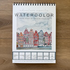 May include: A spiral-bound watercolor workbook with the text "adventure awaits" and "WATERCOLOR WORK BOOK BY KRISTIN DOUGLAS." The cover features a watercolor painting of colorful buildings. Includes 24 ink sketches of bucket list destinations.