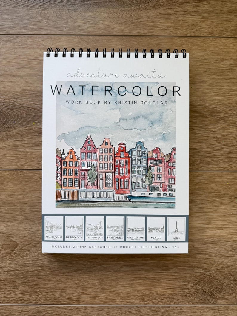 May include: A spiral-bound watercolor workbook with the text "adventure awaits" and "WATERCOLOR WORK BOOK BY KRISTIN DOUGLAS." The cover features a watercolor painting of colorful buildings. Includes 24 ink sketches of bucket list destinations.