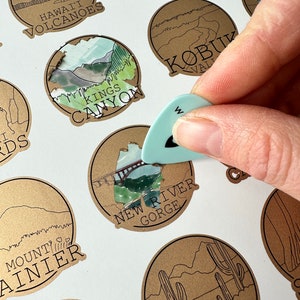 May include: A collection of brown paper circle stickers with illustrations of various national parks. The stickers feature text such as "Hawaii Volcanoes", "Kings Canyon", "New River Gorge", "Mount Rainier", "KOBUK VALLEY", and "SAGUARO".